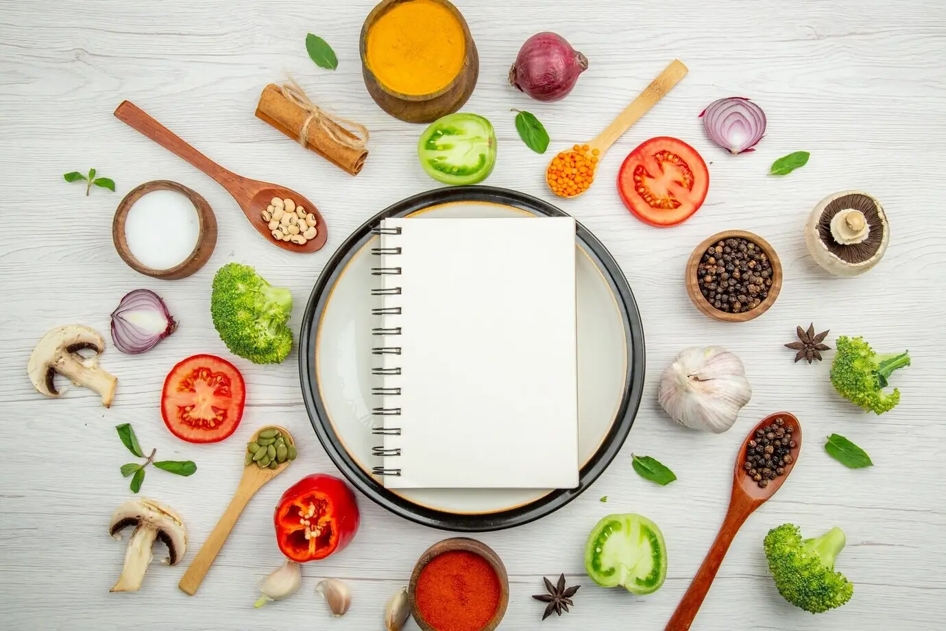 Draufsicht: Notizblock auf weißem Teller, grüne Tomate, schwarzer Pfeffer in einer Schale, Holzlöffel, Knoblauch, Brokkoli und Minzblätter auf grauem Tisch.